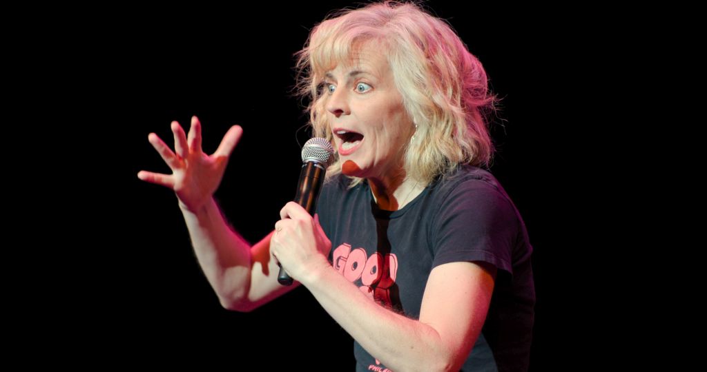 "A woman with blonde hair performs stand-up comedy on stage, holding a microphone and gesturing expressively with one hand, set against a dark background."