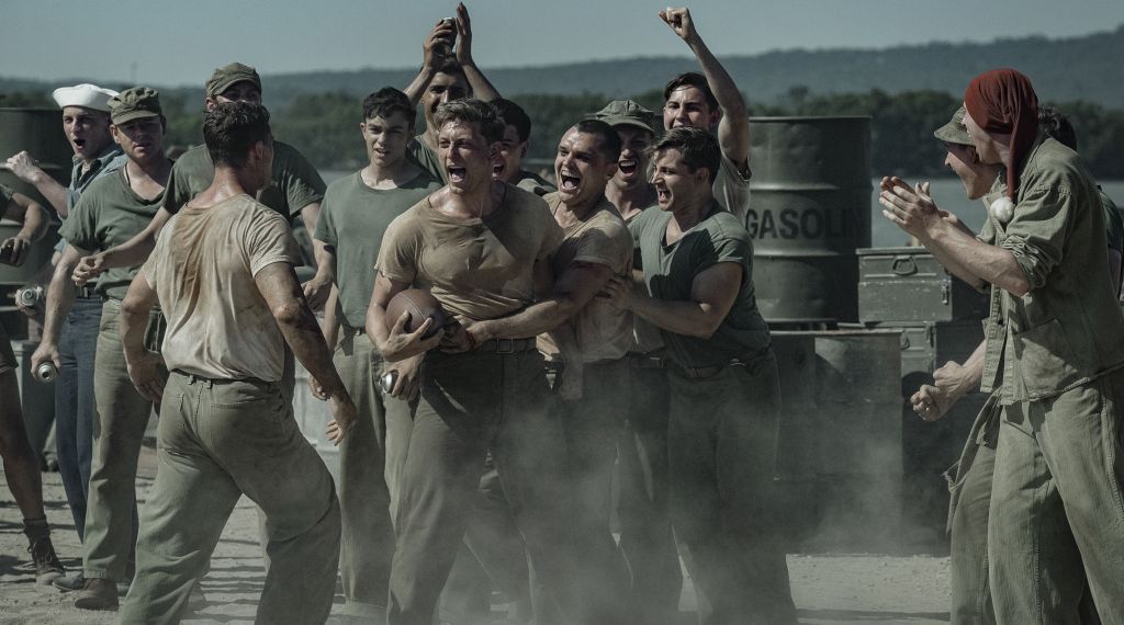"A group of WWII-era soldiers in dusty uniforms are shouting and cheering around two men, possibly during a brawl or contest, outdoors near metal barrels and mountains in the background."
