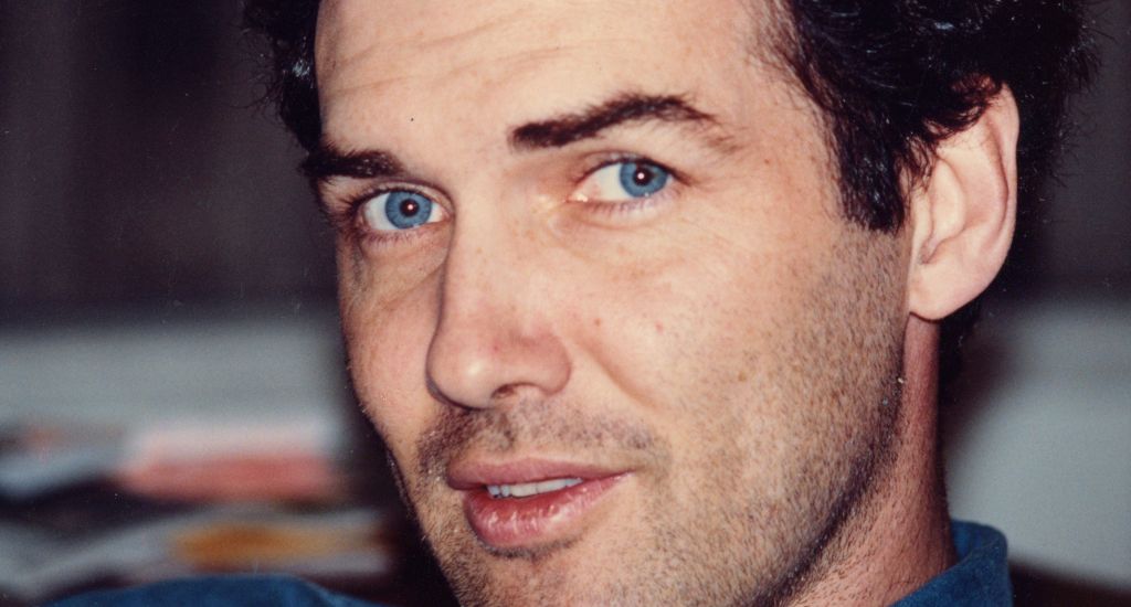 "Man with blue eyes and dark curly hair looking at the camera indoors, with blurred background and casual setting."